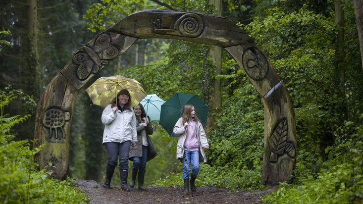 A family wearing raincoats and holding umbrellas walking beneath a wooden archway carved with leaves, snails, butterflies and other nature images at Brockhampton